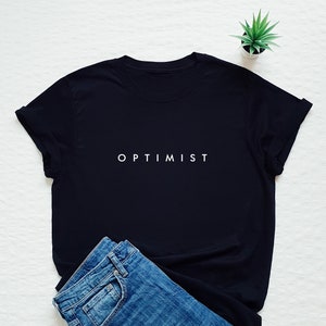Op de afbeelding: Een zwart T-shirt met het woord "OPTIMIST" in wit op de voorkant. Het shirt ligt plat op een wit oppervlak met een blauwe spijkerbroek ernaast.