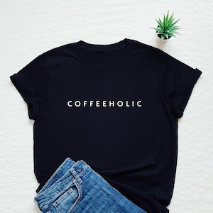 May include: Black t-shirt with white text that reads "COFFEEHOLIC". The shirt is laid flat on a white surface with a pair of blue jeans folded next to it.