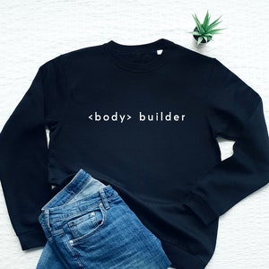Op de afbeelding: Een zwart sweatshirt met witte tekst die "<body> builder" zegt. Het sweatshirt ligt plat op een wit oppervlak met een blauwe spijkerbroek ernaast gevouwen.