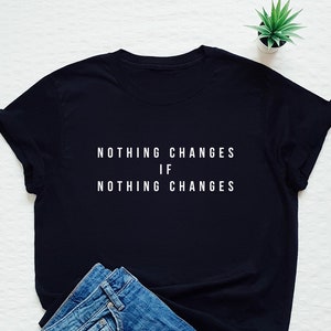 May include: Black t-shirt with white text that reads "Nothing changes if nothing changes". The shirt is laid flat on a white surface with a pair of blue jeans folded next to it.
