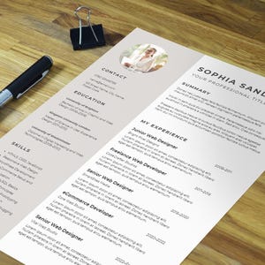 May include: A resume featuring a professional headshot of a woman. The resume is for Sophia Sanders and details her professional experience, education, and skills. The resume is on a light brown wooden surface.