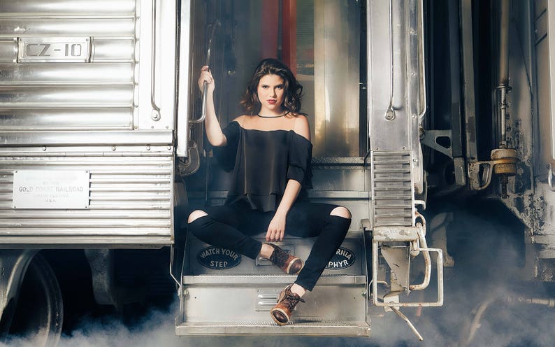 K&ouml;nnte beinhalten: Eine Frau in einem schwarzen, schulterfreien Oberteil und schwarzen Jeans sitzt auf einer Stufe in einem Zugabteil. Das Zugabteil ist aus Metall und hat ein Schild mit der Aufschrift "GOLIATH FREIGHT RAILROAD".