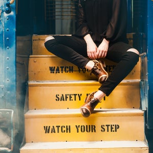 K&ouml;nnte beinhalten: Eine Person in schwarzen Jeans und einem schwarzen Shirt sitzt auf gelben Stufen mit den Worten "Watch Your Step" und "Safety First", die darauf gemalt sind. Die Person tr&auml;gt braune und schwarze Leoparden-Schuhe.