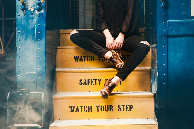K&ouml;nnte beinhalten: Eine Person in schwarzen Jeans und einem schwarzen Shirt sitzt auf gelben Stufen mit den Worten "Watch Your Step" und "Safety First", die darauf gemalt sind. Die Person tr&auml;gt braune und schwarze Leoparden-Schuhe.