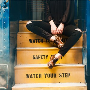 K&ouml;nnte beinhalten: Eine Person in schwarzen Jeans und braunen Lederschuhen mit Leopardenmuster sitzt auf gelben Stufen mit den Worten "Watch Your Step" und "Safety First", die darauf gemalt sind.