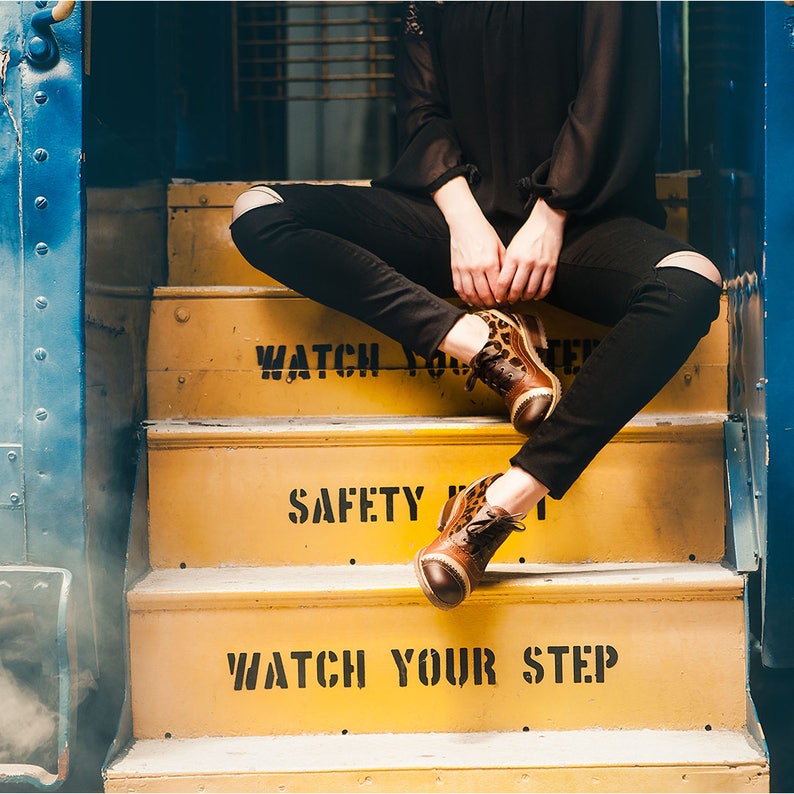 K&ouml;nnte beinhalten: Eine Person in schwarzen Jeans und braunen Lederschuhen mit Leopardenmuster sitzt auf gelben Stufen mit den Worten "Watch Your Step" und "Safety First", die darauf gemalt sind.