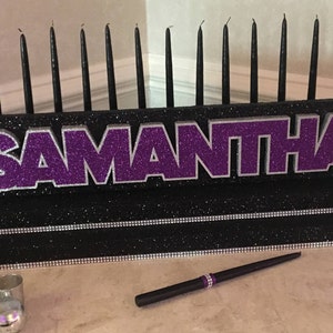 May include: A black candle holder with a purple glitter nameplate that reads "SAMANTHA". The holder has 18 black candles and a black pen with a purple glitter accent.