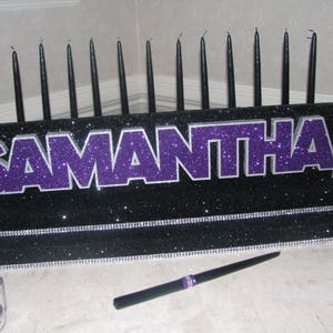 May include: A black and purple glitter sign with the name "Samantha" on it. The sign is decorated with black candles and sits on a black base with a silver glitter trim.