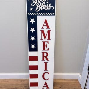 May include: A patriotic wooden sign with a white background and red, white, and blue accents. The sign features the words "God Bless" in white script above "America" in red, capital letters. The design incorporates stars and stripes.