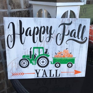 Könnte beinhalten: Ein rechteckiges Holzschild mit weißem Hintergrund und schwarzer Schrift, auf dem "Happy Fall Y'all" steht. Das Schild zeigt einen grünen Traktor, der einen Wagen mit orangefarbenen Kürbissen zieht. Ein oranger Pfeil zeigt nach rechts.