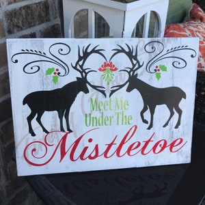 Könnte beinhalten: Rechteckiges Holzschild mit weißem, verwittertem Hintergrund. Zwei schwarze Hirschsilhouetten stehen sich gegenüber, ihre Geweihe bilden ein Herz. Auf dem Schild steht "Meet Me Under The Mistletoe" in grünen und roten Buchstaben, mit Mistel- und Stechpalmenzweigen.