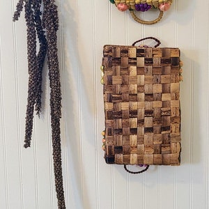 May include: Two woven wall baskets with colorful beads hanging on a white wall. The top basket is round and the bottom basket is rectangular.