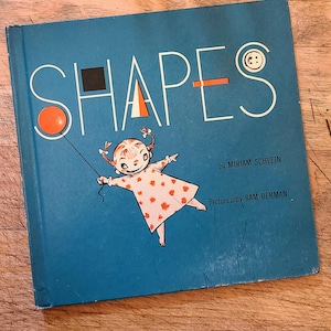 Può includere: Un libro blu con il titolo "SHAPES" in lettere bianche. Il libro presenta un'illustrazione colorata di una ragazza che tiene un palloncino rosso e un piccolo bottone sulla copertina. Il testo "by MIRIAM SCHLEIN" e "Pictures by SAM BERMAN" è stampato sulla copertina.