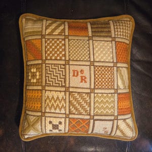 May include: Square decorative pillow with a patchwork design in shades of orange, beige, and brown. The pillow features a grid pattern with various geometric and textured squares. The initials "D&R" are embroidered in the center. The pillow has a gold-colored border.