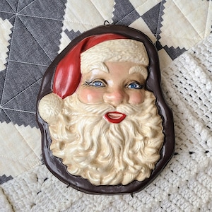 May include: A vintage Santa Claus face ornament with a white beard and a red hat. The ornament is made of plaster and has a brown background.