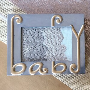 Può includere: Cornice per foto argentata con un'iscrizione 'baby' dorata. La cornice ha una superficie testurizzata e una forma rettangolare.