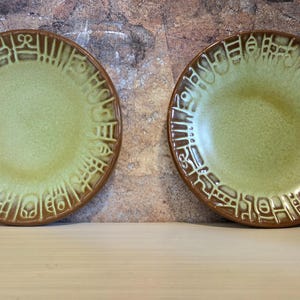 May include: Two round ceramic plates featuring a light green center and a dark brown rim. The rim is decorated with a raised, embossed pattern of abstract geometric shapes and lines that encircle the entire edge of each plate.