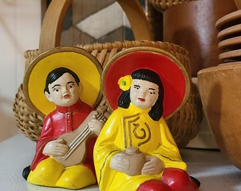 May include: Two ceramic figurines of Asian figures in traditional attire. One figure wears a red outfit and plays a stringed instrument. The other figure wears a yellow outfit and holds a small pot. Both figures wear hats and are seated.