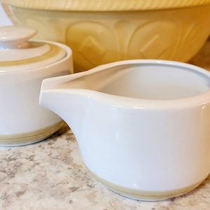 Op de afbeelding: Een witte keramische set van een melkkan en suikerpot met een beige band rond de basis en rand. Een gele mengkom staat op de achtergrond.
