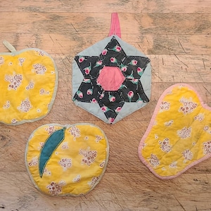 Op de afbeelding: Vier gequilte ovenwanten in de vorm van een appel, een ster en twee peren. De ovenwanten zijn geel met een bloemenpatroon en hebben roze en groene accenten.