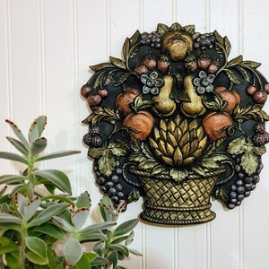 May include: A gold and black wall decoration featuring a basket overflowing with fruit, grapes, and berries. The basket is surrounded by leaves and flowers.