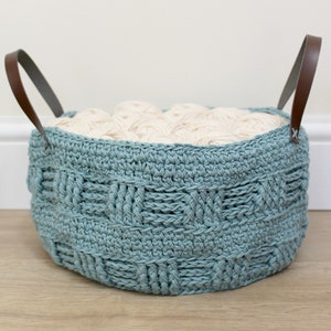 Può includere: Un cesto in crochet color teal con manici in pelle marrone riempito di filato bianco.