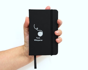 May include: A black notebook with a white yarn ball and the text "Yarn Whisperer" on the cover. The notebook has a black elastic band and a black ribbon bookmark.