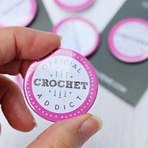 May include: A pink and white round button with the text "OFFICIAL +TTT+ CROCHET +TTT+ ADDICT" in black lettering.