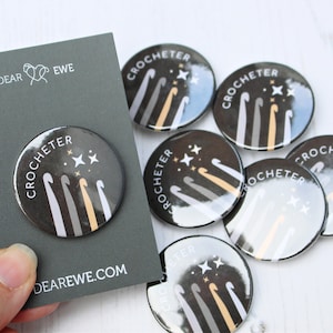 May include: A set of six black and white round buttons with the text "Crocheter" and a graphic of crochet hooks and stars. The buttons are on a white surface.