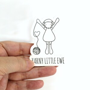 May include: Black and white sticker of a sheep holding a ball of yarn with the text "Yarn Little Ewe" below.