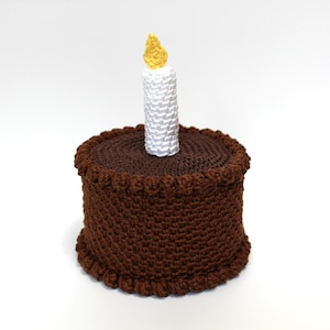 May include: A brown crocheted cake with a white crocheted candle on top. The candle has a yellow flame.