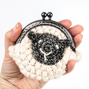 May include: A crocheted coin purse shaped like a sheep with a black face and white wool. The purse has a metal clasp.