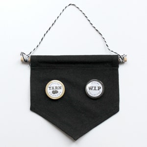 May include: Black fabric banner with a white string hanging from wooden dowels at the top. Two round pins are attached to the banner. The left pin is gold with the text "OFFICIAL YARN SQUISHER" and the right pin is silver with the text "OFFICIAL W.I.P. COLLECTOR".