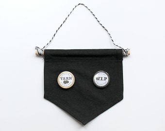 May include: Black fabric banner with a white string hanging from wooden dowels at the top. Two round pins are attached to the banner. The left pin is gold with the text "OFFICIAL YARN SQUISHER" and the right pin is silver with the text "OFFICIAL W.I.P. COLLECTOR".