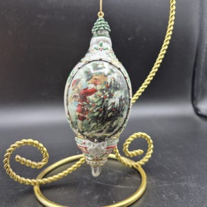 May include: An oval Christmas ornament with a painted scene of a person in a red coat decorating a Christmas tree. The ornament has gold trim and is suspended from a gold chain. The ornament is displayed on a gold metal stand.