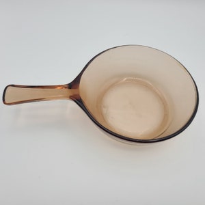 May include: A vintage amber-colored glass saucepan with a handle. The pan has a rounded shape and a dark brown rim. The handle is also amber-colored and has a small logo. The saucepan is empty and sits on a white surface.