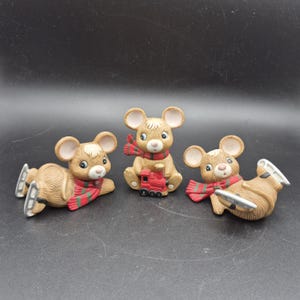 May include: Three ceramic mouse figurines. Each mouse is light brown with large ears, wearing a red and green scarf. One mouse is sitting with a red train, and two are wearing ice skates.