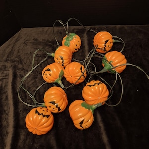 May include: A string of ten orange plastic jack-o'-lantern light covers connected by a dark green electrical cord. Each pumpkin features carved facial expressions with black painted details and green plastic stems at the top of each individual light.