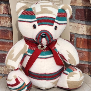 May include: A white teddy bear with a red, green and brown patterned fabric. The bear is wearing a red ribbon with a silver charm around its neck.