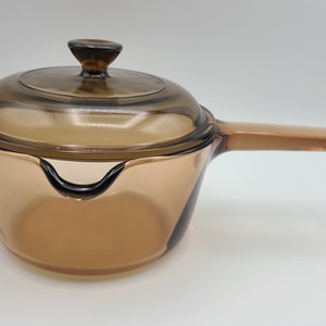 May include: A clear, amber-colored saucepan with a matching lid. The saucepan has a long handle and a pouring spout. The lid has a small knob. The cookware is made of glass and is designed for stovetop use.