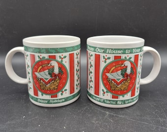 Puede incluir: Dos tazas de cerámica blanca con temática navideña. Cada taza presenta un diseño a rayas rojas y blancas con una imagen circular de un ganso en una cesta. También se ven acebo verde y las palabras "Happy Holidays".