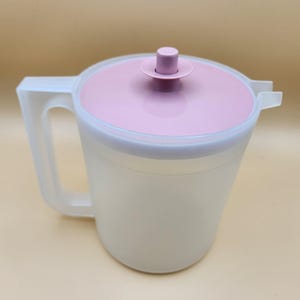 Può includere: Una caraffa di plastica trasparente con un coperchio rosa e un manico. Il coperchio ha un piccolo pomo rosa in cima.