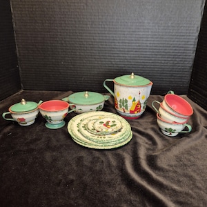 May include: A vintage metal toy tea set featuring a white teapot with a green lid and farm scene, a lidded sugar bowl, a creamer, stacked plates with a rooster motif, and several cups with pink interiors and green exterior trim details.