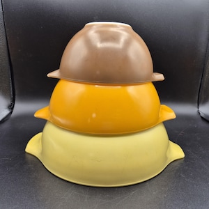 May include: Three vintage Pyrex mixing bowls stacked on top of each other. The bowls are in shades of brown, orange, and yellow. Each bowl has a handle on either side.