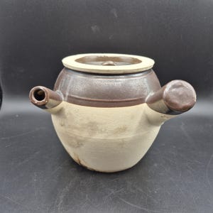 May include: A ceramic pot with a lid, featuring a two-tone design. The upper section and the lid are a dark brown, while the lower portion is a creamy white. It has two spouts and a handle on the lid. The pot is likely used for cooking or serving.