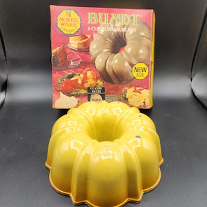 Puede incluir: Un molde amarillo para bizcocho Bundt con un diseño acanalado, junto a una caja con el texto "BUNDT" y "6 CUP FLUTED TUBE PAN". La caja presenta imágenes de productos horneados y las palabras "NORDIC WARE" y "NEW SIZE".