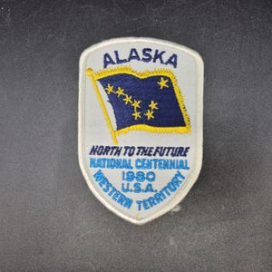 May include: A shield-shaped embroidered patch with a light gray background. It features a dark blue flag with yellow stars, the text ALASKA, NORTH TO THE FUTURE, NATIONAL CENTENNIAL, 1980, U.S.A., and WESTERN TERRITORY in blue and yellow thread.
