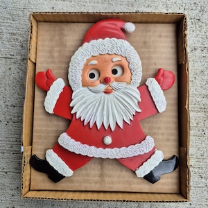May include: A molded plastic Santa Claus wall decoration featuring a red suit with white textured trim, a matching red hat, black boots, and red mittens. The figure has a white beard, a peach-colored face, and is presented inside a cardboard box.