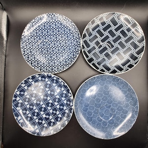 May include: Four round ceramic plates arranged in a square formation. Each plate features a distinct dark blue and white pattern: one with a diamond grid, one with a rectangular weave, one with a cross-hatched geometric design, and one with a spiral motif.