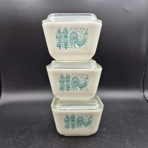 Könnte beinhalten: Drei gestapelte, cremefarbene Pyrex-Vorratsbehälter mit Deckeln aus klarem Glas. Jeder Behälter hat ein tealfarbenes Hahn- und Blumendesign. Die Behälter sind quadratisch und scheinen Vintage zu sein.